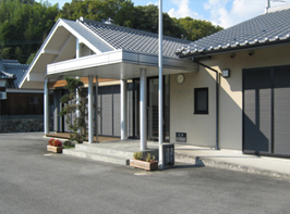 青空の下後ろに青々とした木々の映る、白い外観と灰色の瓦屋根の尾崎公民館の写真