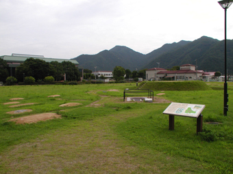 緑色の屋根の大きな施設の手前に、中央に円形の古墳のような物や案内看板が広大な敷地内に設置された新宮宮内遺跡の写真