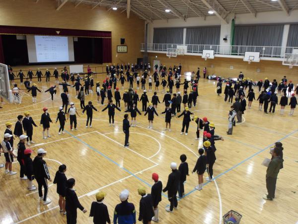 体育館で円形に並んだ児童たちが中心にいる男性の説明を聞いている写真