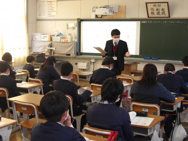 黒板の前に立って話をしている先生を生徒たちが真剣に聞いている授業の様子を後方から写した写真