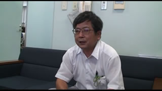 横長の椅子に座った白いシャツを着た男性担当者がインタビューされている様子をアップで撮影した写真