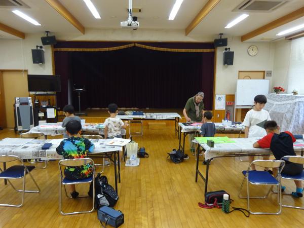 前方に舞台がある室内で、児童達が長机に新聞紙を広げ絵を描いている写真