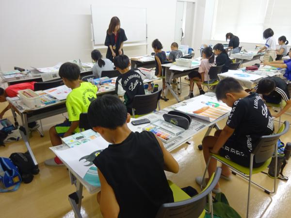 前方にホワイトボードが置かれた室内で、児童達が長机に2名ずつ座り絵の具セットを広げ白い画用紙に絵を描いている写真