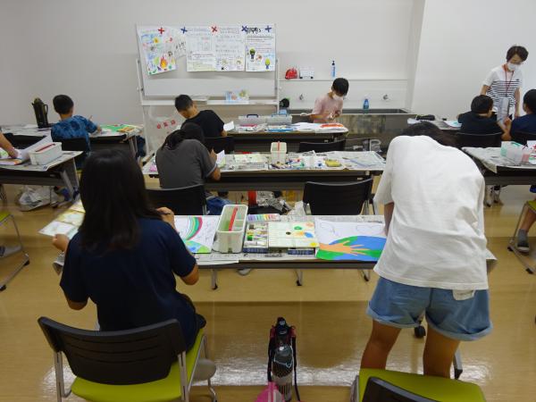 児童達が長机に2人ずつ座り、絵の具セットを広げ白い画用紙に絵を描いている写真
