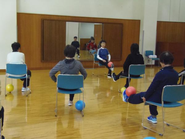 椅子に座った講師の前に5人程の参加者が椅子に座り左足をまっすぐに伸ばし足の先でボールを載せている人や足だけまっすぐに伸ばしているチェアビクスの写真