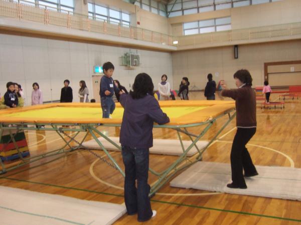 大きな黄色のトランポリンの上を幼児が歩きトランポリンの左側で手を差し伸べている保護者と前で待っている講師の親子体操の写真
