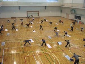 体育館で多くの人が両足を広げ、まっすぐ立ち上半身を前に曲げ両手を広げている健康レクリエーションの写真
