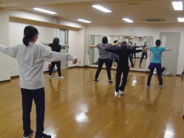 鏡の前に指導者が立ちダンスの見本を見せ5人程の参加者が立って両手を広げているリズムダンスの写真
