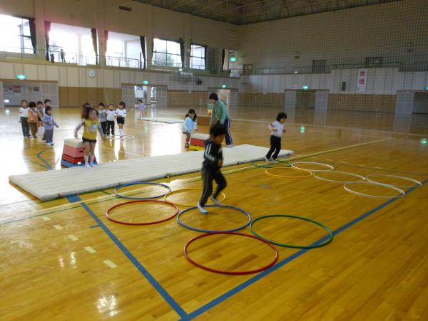 体育館にとび箱、マット、フラフープを横に倒し5つ並べたものが2列あり、とび箱の前で順番待ちしている幼児、とび箱を跳んだ幼児、フラフープの上走っている幼児ののびっ子体操教室の様子の写真