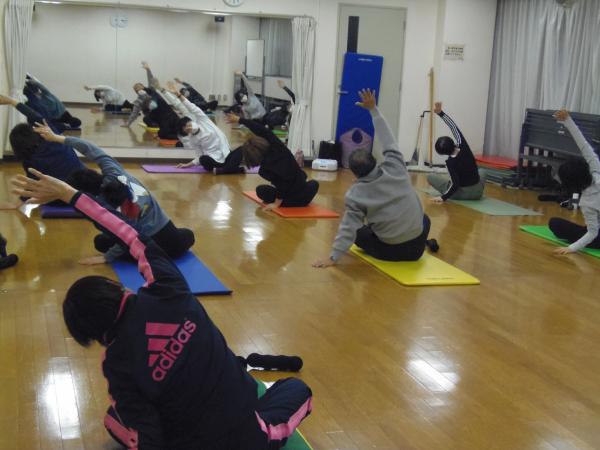 鏡の前に座ってポーズをしている講師の前に7人程の参加者があぐらで座り右手を上げ左側に上体を曲げているヨガの写真