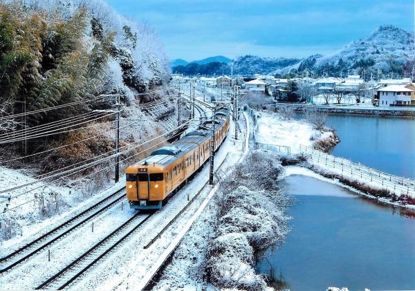 雪化粧をしている山々を背景に、雪が降り積もる川沿いの線路をオレンジ色の電車が走っている写真