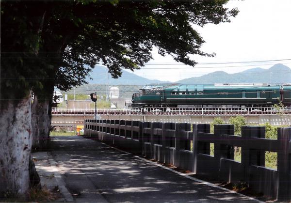 石の柵が並んでいる小道の先の線路を緑色の列車が横切っている写真