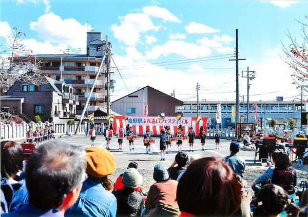よく晴れた青空の下、竜野駅ふれあいフェスティバルと書かれた幕が張られた広場でマーチングをしている子どもたちと、それを見ている観客たちの写真