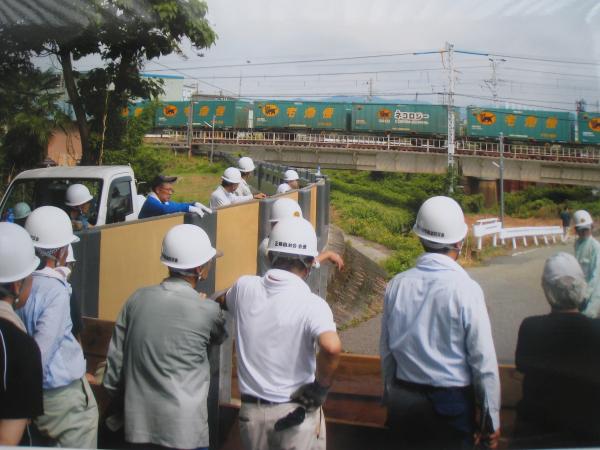 白いヘルメットを被った大勢の大人たちの奥を走る貨物列車を撮影した写真