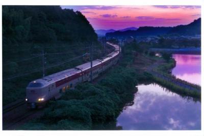 夕日が暮れている池の横をライトを点けて走る電車の写真