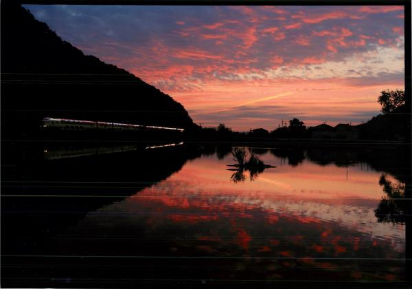 夕焼けの空が鏡のように反射する水面の横を走り抜ける電車を撮影した写真