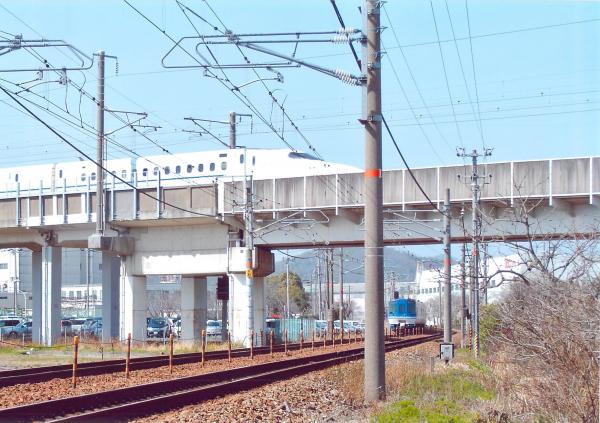 線路を走る青い電車の上に掛かる鉄道橋を早い速度で走っていく新幹線を撮影した写真