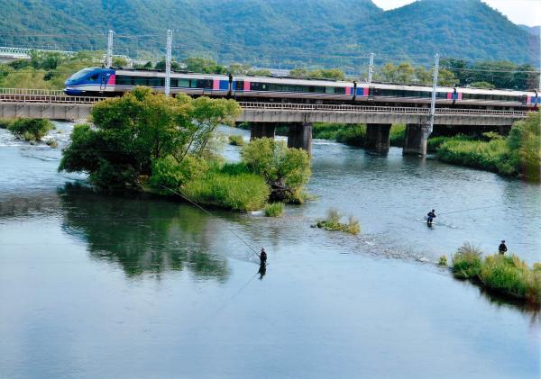 渓流釣りをする釣り人の近くにある橋の上の線路を颯爽と走る青い電車を撮影した写真