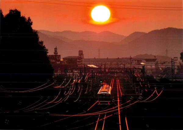 オレンジ色に光る夕日をバックに駅から発進する電車を撮影した写真