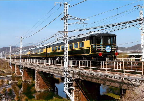 流れが緩やかな川の上の橋を走っている黄色と黒の電車を撮影した写真