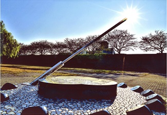 公園の広場のような場所でハートの台座に寄り掛かった銀色のアイスピックのようなオブジェの頂点に太陽が重なった瞬間の写真