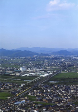青空の下所々に緑の畑がある市街地の中にある線路を、ドクターイエローと呼ばれる黄色の新幹線が通過している様子を上空から撮影した写真