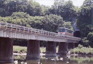 木々の生えた山のトンネルを抜けた赤いラインの入った1両の電車が、川にの上の橋を渡っている写真