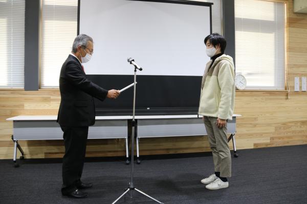 みんなの前で最優秀賞の賞状を授与される学生さんと、その賞状を読み上げるスーツ姿の男性を横から写した写真
