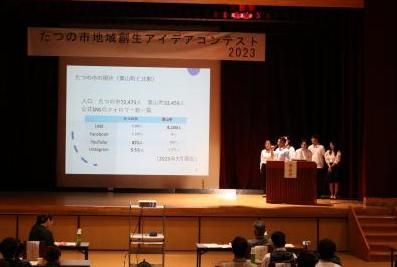 舞台の上に立ち大きなスクリーンに映される資料とともに政策提言を行う学生さんたちの写真