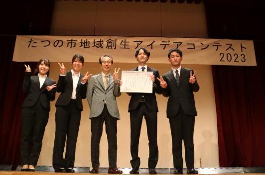 最優秀賞を受賞した同志社大学田中ゼミAチームの皆さんが、舞台の上に立ち賞状を持ってピースサインをしている写真
