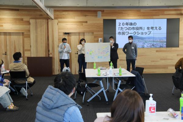 4名の参加者が、たくさんの付箋が貼られた大きな模造紙を持ってみんなの前で発表をしているような様子を写した写真