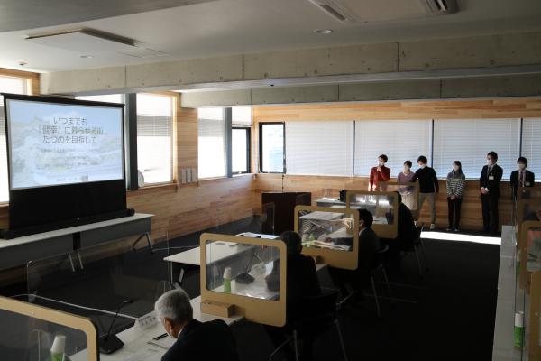 広い部屋で大きなモニターに映された資料を見ながら発表をする学生さんたちと、その発表を長机の席に座って聴くスーツ姿の人々を写した写真