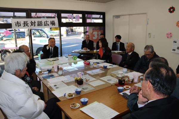 「市長対話広場」と書かれた紙がガラス戸に貼られた室内で参加者達がテーブルの席に座り、市長対話広場が行われている写真