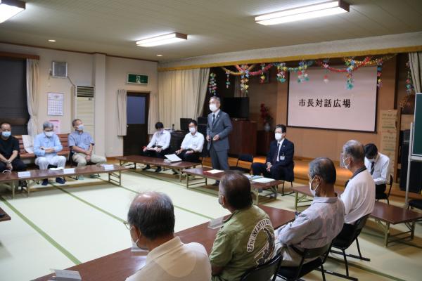 畳が敷かれた広い室内に設置されている座卓タイプの長机に参加者達が座り、室内前方で男性が立ち話をしている写真