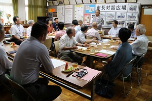 15名前後の参加者達が席に座り、前方で話している男性の話を聞いている様子の写真