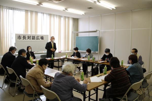 「市長対話広場」と書かれた紙がホワイトボードに貼られ、ロの字型に設置されたテーブルの席に参加者達が座り、市長対話広場が行われている写真