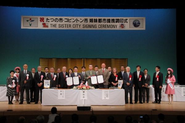 「たつの市・コビントン市姉妹都市提携調印式」と書かれた幕の下のステージ上に多くの人が横一列に並んで撮影した写真