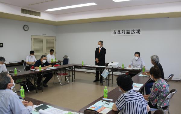ロの字型に設置された机の席に参加者達が座り、1人の男性が手を前に組んで立って話をしている写真