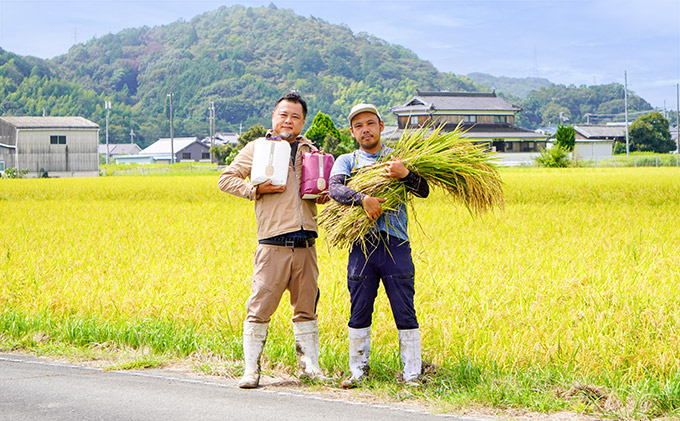 収穫時の田んぼを背景に米袋と稲穂を抱えて立つ2人の男性の写真