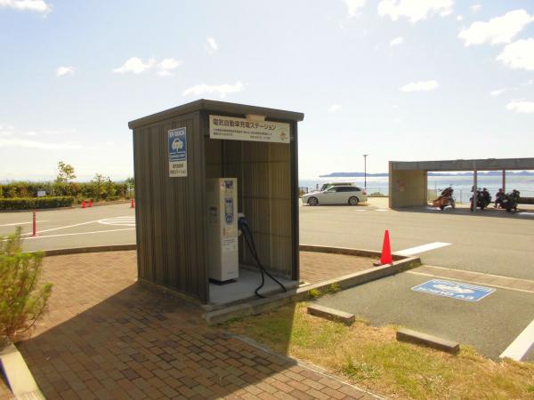 海を背景に、駐車場の脇の小屋の中に設置されている電気自動車用急速充電器の写真