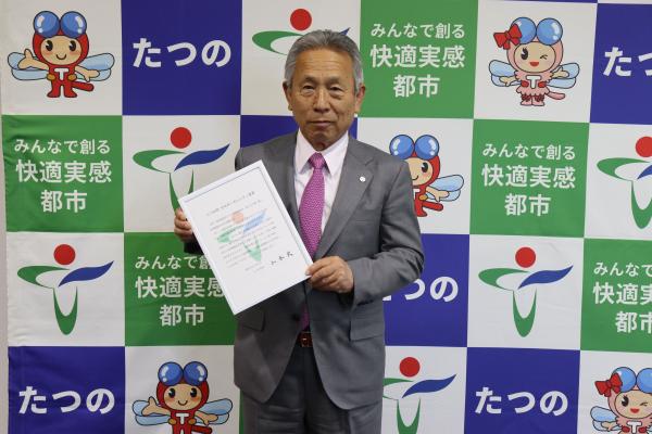 青緑白の竜野市のバックパネルの前で、グレーのスーツを着た竜野市長が書類を手に持ち正面を向いている写真