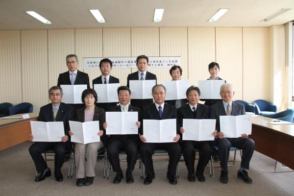 会議室で複数のスーツなどを着た人たちが、協定書を手に持ち並んでこちらを見ている写真