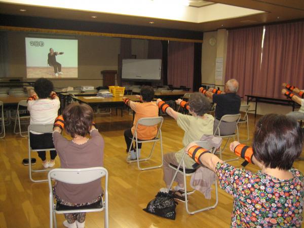 いきいき百歳体操で室内前方のスクリーンに映し出された体操の映像と同じ動きをしている参加者達の写真