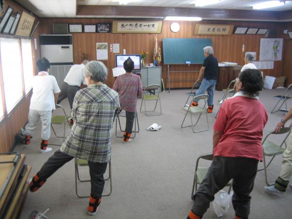 参加者達がパイプ椅子の後ろに立ち、背もたれ部分を支えにしながら左足を横に広げ、片足立ちしている写真
