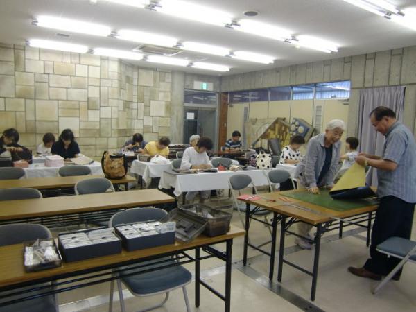 革細工体験教室の参加者たちが、縦二列に置かれた長机の席に座って革細工作りをしている写真