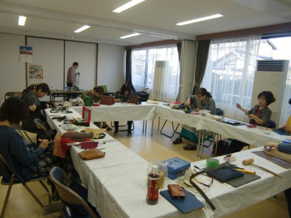 革細工体験教室の参加者たちが、ロの字形に配置された長机の席に座って集中して革細工作りをしている写真