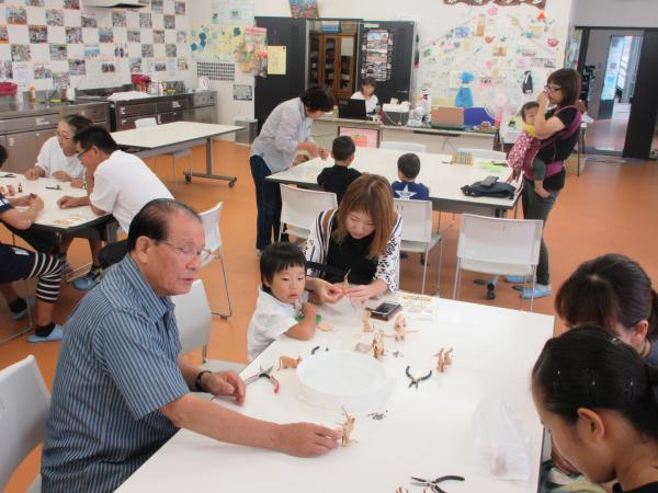 革細工の体験教室に参加した大人たちや子どもたちが、広い部屋で一緒に革細工作りをしている写真