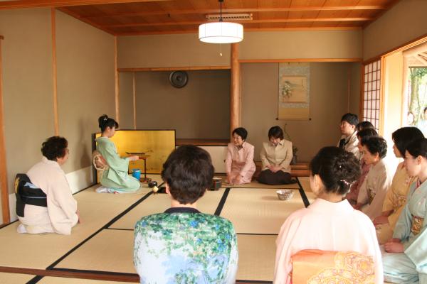 着物やスーツを着た女性たちが、楽庵の広間に正座してお茶会を開いている様子を後方から写した写真