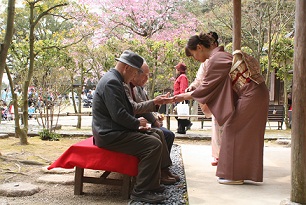 聚遠亭の敷地内に置かれた長椅子に腰かける男性たちに、何かを手渡しする着物姿の女性たちの写真