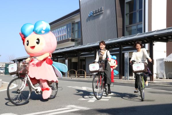 たつの市のイメージキャラクターのあかねちゃんと二人の女性が、本竜野駅前のロータリーをレンタル自転車に乗って走っている写真